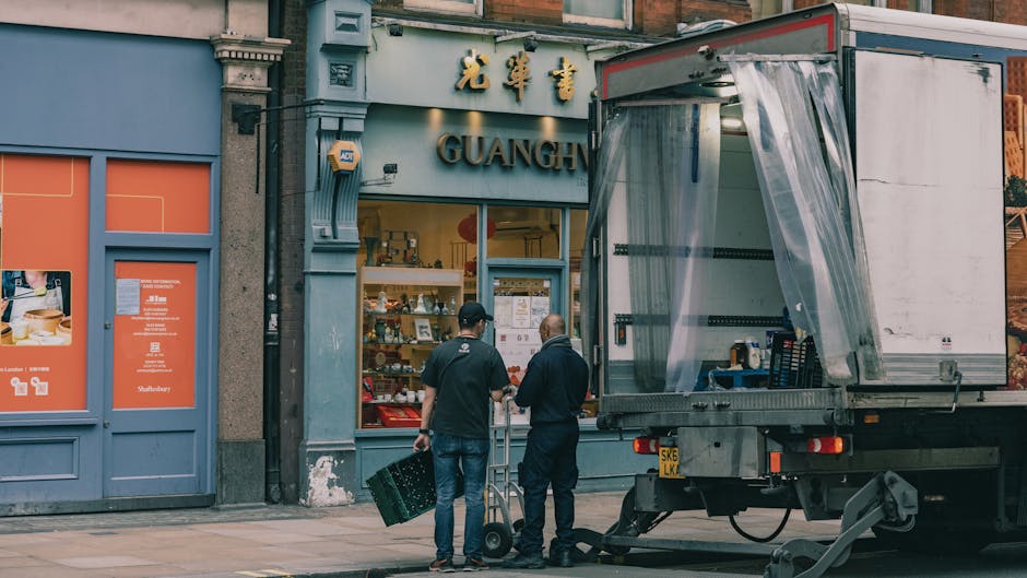 Two workers are engaged in a home relocation process outside a store named GUANGH. One worker, wearing a black cap and dark clothes, holds a black crate in one hand and is standing next to a hand trol