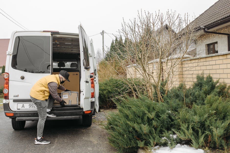 A man wearing a yellow padded jacket, grey trousers, a black cap, and gloves is engaged in loading cardboard boxes into the rear compartment of a white moving van parked outside a residential property. The van's rear doors are open, revealing several packed boxes placed on the vehicle's cargo area floor. The man is bending forward, holding a box in both hands, preparing to lift or place it inside the van. Nearby, in the driveway, there are additional packed boxes and packing materials such as plastic wrap and tissue paper. The property features an exterior brick wall and a window, with a roof that has visible string lights hanging along the eaves. To the right of the van, there are leafless shrubs and evergreen plants, indicating a garden area. The scene is set during daytime with overcast weather, illustrating a home relocation process involving furniture transport, packing, and loading activities often associated with professional removals services like those offered by Man with Van Fulham.