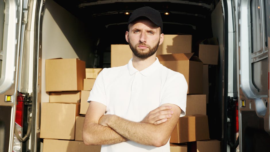 A young man with a beard, wearing a black cap and a white polo shirt, stands with arms crossed inside the back of a loading van filled with packed cardboard boxes of various sizes, some wrapped in plastic or fabric. The van is parked on a paved area, with the open rear doors revealing the interior environment. The boxes are positioned in the cargo space, indicating an organized packing process typical of house removals or furniture transport. The lighting inside the van highlights the boxes and the man, suggesting daylight. This scene reflects aspects of home relocation, packing, and logistics handled by a moving service such as Man with Van Fulham, specializing in removals and furniture transport.