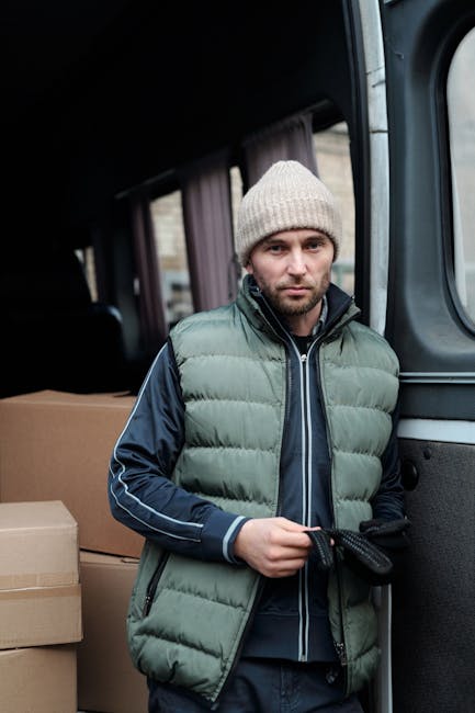 A man wearing a yellow padded jacket, grey trousers, a black cap, and gloves is engaged in loading cardboard boxes into the rear compartment of a white moving van parked outside a residential property. The van's rear doors are open, revealing several packed boxes placed on the vehicle's cargo area floor. The man is bending forward, holding a box in both hands, preparing to lift or place it inside the van. Nearby, in the driveway, there are additional packed boxes and packing materials such as plastic wrap and tissue paper. The property features an exterior brick wall and a window, with a roof that has visible string lights hanging along the eaves. To the right of the van, there are leafless shrubs and evergreen plants, indicating a garden area. The scene is set during daytime with overcast weather, illustrating a home relocation process involving furniture transport, packing, and loading activities often associated with professional removals services like those offered by Man with Van Fulham.