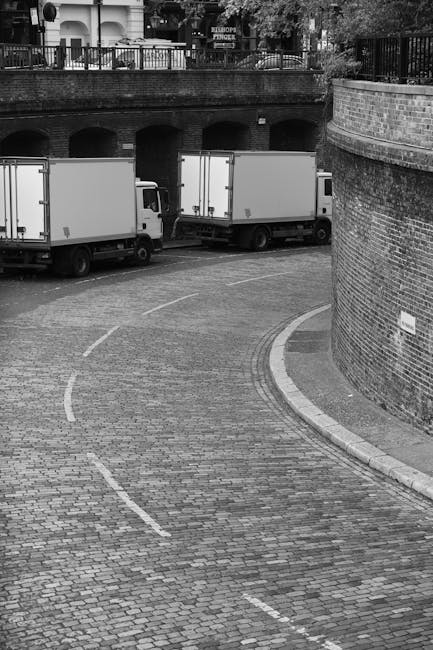 A black-and-white photograph showing a curved cobblestone driveway leading to a loading area beneath a brick bridge structure. Two white moving trucks are parked side by side on the driveway, with the rear ends facing the overhead bridge. The trucks are positioned to facilitate a home relocation process, likely involving the transfer of furniture, boxes, and appliances. The street is lined with a stone curb and a brick wall curves along the right side of the image, with a small sign attached to it. Above the bridge, glimpses of residential buildings and greenery are visible, with some fencing and trees partly obscured by the bridge structure. The scene appears to be part of a professional removal or moving operation, as coordinated by companies such as Man with Van Fulham, highlighting the logistics involved in house removal services on New Kings Road to Fulham Palace.