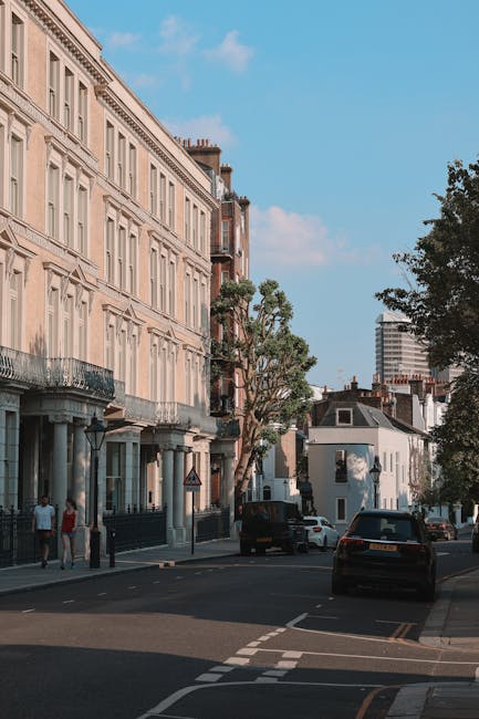 A residential street scene in Fulham with a row of three to four-storey Victorian-style terraced buildings featuring cream and light brown facades, decorative moldings, and black iron balconies. Several large, leafy trees line the pavement, casting shadows on the street. Parked cars, including a black van, are visible along the curb. Pedestrians are walking on the sidewalk, and a portable traffic sign is positioned at the corner. The sky is mostly clear with a few clouds, and a modern high-rise building is visible in the background. This scene represents an urban environment suitable for house removals and furniture transport services by Man with Van Fulham, highlighting the typical setting for home relocation logistics and moving processes.
