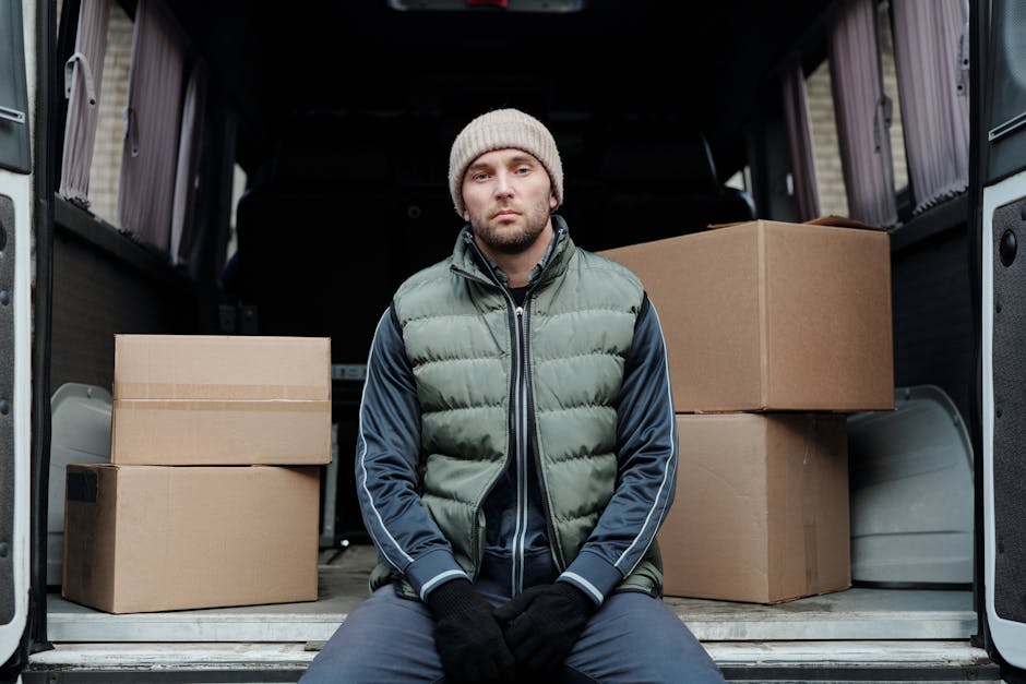 A man wearing a black vest, dark pants, and a baseball cap is inside the rear of a white moving van, which is parked on a paved area outdoors under an overcast sky. He is in the process of loading or 