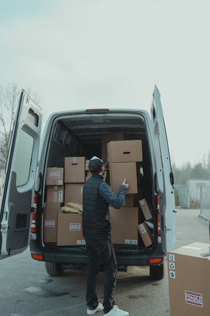 A man wearing a dark jacket, jeans, and a black cap is standing at the open rear doors of a white moving van, inside which numerous cardboard boxes of various sizes are stacked. Some boxes have printed labels or barcodes, and others appear to be plain. Several boxes are placed on the floor of the van, while others are arranged on top of each other, indicating an organized packing process typical of home relocations. The man is holding a box on his right side, possibly preparing to load it onto or unload it from the vehicle. The van is parked on a paved surface, with a cloudy sky overhead and a blurred background of trees and industrial buildings, reflecting an outdoor moving or logistics scene. The environment suggests a professional furniture transport or packing and moving service, aligned with house removals in the Fulham area, as provided by Man with Van Fulham.