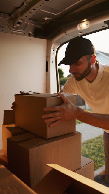 A man wearing a black cap and a cream-colored shirt is inside the back of a moving van, engaged in loading or arranging cardboard moving boxes. The boxes are stacked on the van's interior floor and shelf, with some being held by the man while others are placed nearby. The van's interior features a light-colored ceiling with visible fixtures and a window showing a blurred outdoor background of trees and sky. The scene depicts a professional home relocation process, with the man carefully handling packing materials in preparation for a furniture transport or house move, reflecting the services of Man with Van Fulham specializing in removals and moving logistics.