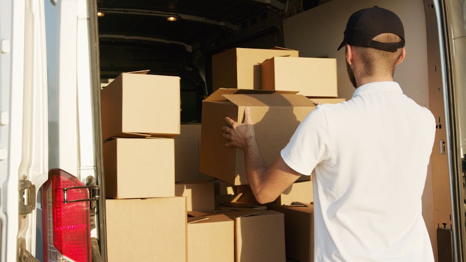 A man wearing a dark jacket, jeans, and a black cap is standing at the open rear doors of a white moving van, inside which numerous cardboard boxes of various sizes are stacked. Some boxes have printed labels or barcodes, and others appear to be plain. Several boxes are placed on the floor of the van, while others are arranged on top of each other, indicating an organized packing process typical of home relocations. The man is holding a box on his right side, possibly preparing to load it onto or unload it from the vehicle. The van is parked on a paved surface, with a cloudy sky overhead and a blurred background of trees and industrial buildings, reflecting an outdoor moving or logistics scene. The environment suggests a professional furniture transport or packing and moving service, aligned with house removals in the Fulham area, as provided by Man with Van Fulham.