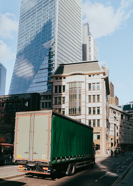 A large green shipping container is positioned on the street beside a black and orange delivery van, situated in an urban environment surrounded by high-rise office buildings and older architectural s